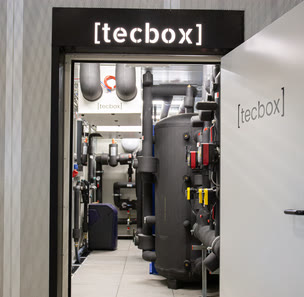 Offene Tür einer [tecbox] mit beleuchtetem Schriftzug, dahinter Speicher und Verrohrung.